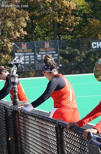 A group of athletes wearing orange uniforms are on the sideline of a sports field, resting against a fence. Behind them, a large digital scoreboard displays team names and scores. The field is bright green, and several banners with sponsor logos and championship wins are visible. The athletes appear to be taking a break, possibly before or during a field hockey game.