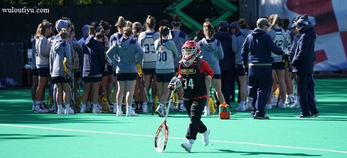 A sports team gathers on a field with a coach, all dressed in athletic gear, while one player wearing a red helmet and jersey walks away from the group carrying a lacrosse stick. The field is green, and there are sports equipment and water bottles visible.