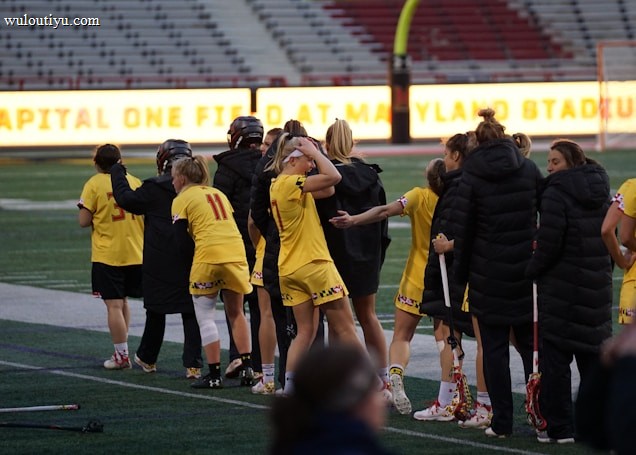 A group of athletes wearing yellow sports uniforms are standing on a sports field. Some are holding lacrosse sticks, and they appear to be interacting casually. Several people are dressed in black coats. In the background, there is a bright sign with text and parts of a stadium are visible.