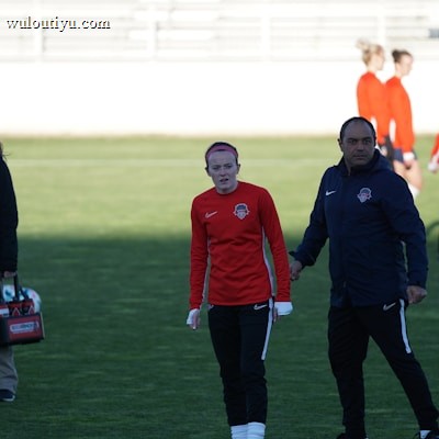 A group of people are gathered on a sports field. Some individuals are dressed in athletic gear, including a woman in a red sports jersey and a man beside her in a blue jacket, appearing to be in conversation. Additional people in similar athletic clothing are visible in the background.