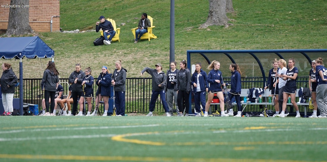 A group of female athletes, some in sports uniforms and others in jackets, are gathered along the sideline of a sports field. They seem to be in a team environment, possibly preparing for or observing a game. In the background, there are a few people sitting on yellow chairs on a grassy incline, and a building with a brick facade.