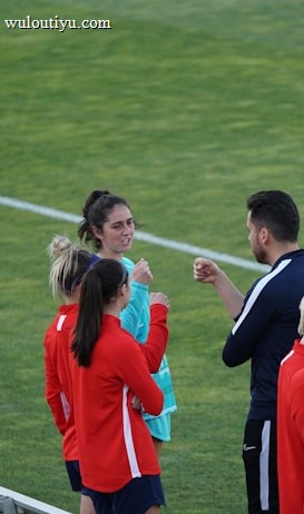 A group of people wearing sports attire are gathered on a grassy field. Some individuals are dressed in red jackets, while others are in turquoise and navy blue. A coach appears to be giving instructions. Sports equipment, such as water bottles and a bench with towels, can be seen nearby.