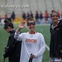 Several athletes in sports attire are on an outdoor sports field, interacting and smiling. One individual in the foreground waves with a friendly expression, while another stands beside her, also smiling. They wear protective goggles and gear associated with lacrosse. The background features indistinct figures in dark clothing against a backdrop of stadium seating.