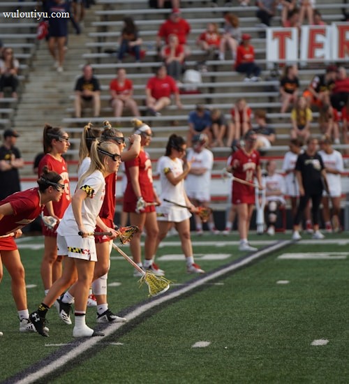 Athletes are lined up on a sports field, preparing for a game. They are dressed in athletic uniforms with some in white and others in red. The players hold lacrosse sticks and wear eye protection. In the background, spectators fill the stadium seats, mostly dressed in casual attire. A large banner is displayed behind them.