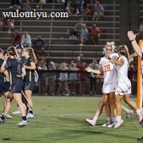 Players wearing sports uniforms are gathered on a lacrosse field. A team in dark uniforms and a team in white uniforms with one player wearing number 28 are seen celebrating near a goalpost. A referee in a striped shirt is raising an arm, indicating a call or score. Spectators are visible in the stands behind.