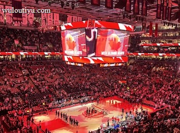 An indoor sports arena filled with spectators and a basketball court illuminated by red lighting. The scoreboards and video screens display promotional content, with numerous banners and championship flags hanging from the ceiling. Players and officials are gathered on the court, possibly in preparation for a game or event.
