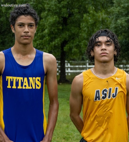 Two young athletes wearing sleeveless sports tops stand on a grassy field. The athlete on the left is wearing a blue top with the word 'TITANS' while the one on the right wears a yellow top with 'ASIJ'. The background features green trees and a white fence, with a bench visible on the right.